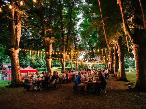 Menschen sitzen an langen Tischen unter Lichterketten im schattigen Schlosspark Kaarz bei einer festlichen Abendveranstaltung im Sommer.